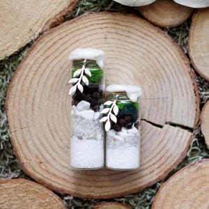 May include: Two small glass jars filled with layers of white, clear, and dark brown materials, topped with green moss and white wax. Each jar has a silver leaf charm and is set on a wooden slice.