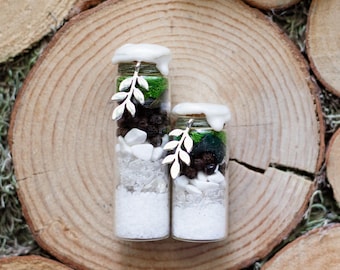 May include: Two small glass jars filled with layers of white, clear, and dark brown materials, topped with green moss and white wax. Each jar has a silver leaf charm and is set on a wooden slice.