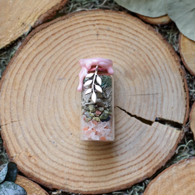 May include: A small glass bottle filled with layers of colorful stones, greenery, and crystals. The bottle is sealed with pink wax and a silver leaf charm. The bottle is displayed on a wooden surface.