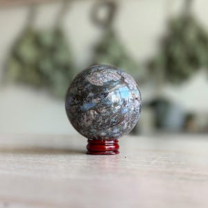 Könnte beinhalten: Eine blaue und graue Kugel mit einem wirbelnden Muster, die auf einem roten Holzständer ruht. Die Kugel ist aus einem Naturstein, möglicherweise einer Art Achat.