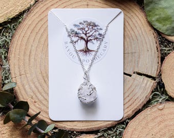 May include: A silver-toned necklace with a pendant featuring a light-colored, irregularly shaped stone wrapped in wire. The pendant hangs from a delicate chain. The necklace is displayed on a white card with the text "BAYOU APOTHECARY" and a tree graphic.