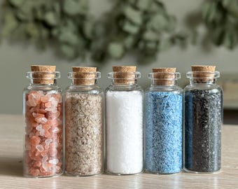 Puede incluir: Cinco pequeños frascos de vidrio con tapas de corcho llenos de sales de diferentes colores. Las sales son rosadas, beige, blancas, azules y negras.