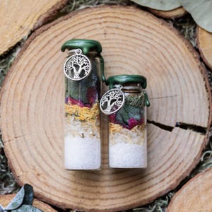 May include: Two glass bottles filled with layers of white crystals, dried herbs, and flower petals. Each bottle has a green wax seal and a silver tree of life charm. The bottles are displayed on a wooden surface.
