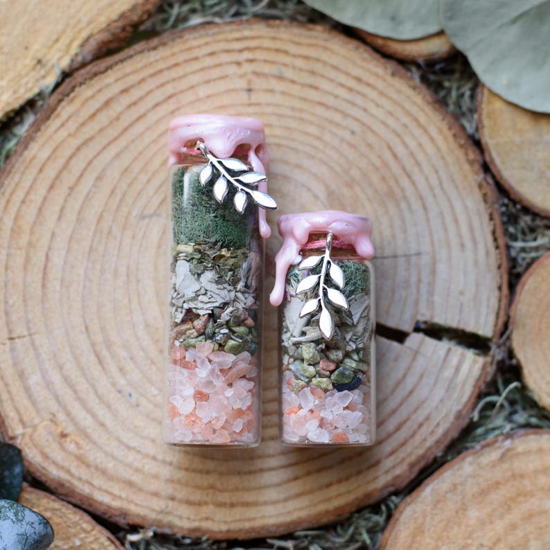 May include: Two glass bottles filled with layers of colorful ingredients, including pink crystals, green herbs, and other natural elements. The bottles are sealed with pink wax and adorned with silver leaf charms. The bottles are displayed on a wooden surface.