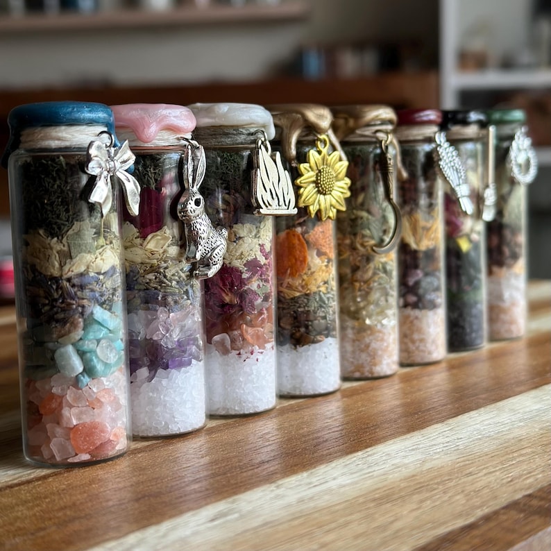May include: A row of eight clear glass cylindrical bottles filled with layered botanical materials, crystals, and salts. Each bottle is sealed with colored wax and features a unique metal charm, including a bow, rabbit, flame, and sunflower.