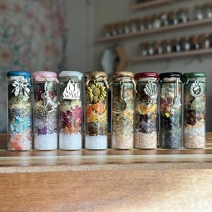 May include: Eight clear glass vials filled with layered dried herbs, flower petals, and colorful crystals. Each vial is sealed with colored wax and features a silver or gold-toned metal charm, including a bow, rabbit, flame, sunflower, and crescent moon.
