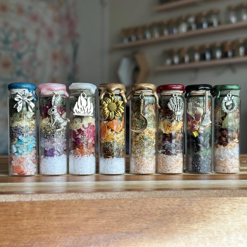 May include: Eight clear glass vials filled with layered dried herbs, flower petals, and colorful crystals. Each vial is sealed with colored wax and features a silver or gold-toned metal charm, including a bow, rabbit, flame, sunflower, and crescent moon.