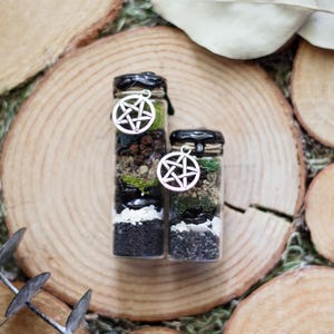 May include: Two small glass bottles filled with layers of black, white, and green materials, topped with black wax and a silver pentagram charm. The bottles are displayed on a wooden slice.
