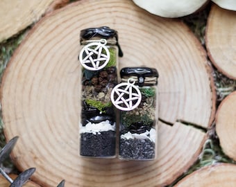May include: Two small glass bottles filled with layers of black, white, and green materials, topped with black wax and a silver pentagram charm. The bottles are displayed on a wooden slice.
