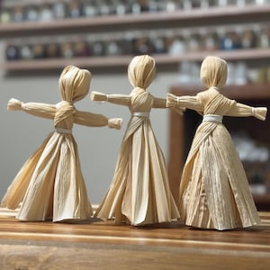 May include: Three handcrafted dolls made from dried corn husks, each featuring a tied waist with white string, outstretched arms, and a full, flared skirt. The figures are light tan in colour, showcasing the natural texture and fibrous detail of the husks.