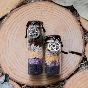May include: Two glass bottles filled with layers of black, purple, tan, and green materials. Each bottle has a black wax seal and a silver pentacle charm. The bottles are displayed on a wooden surface.