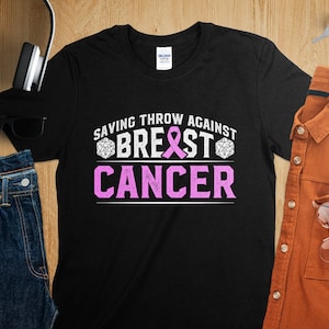 Op de afbeelding: Zwart T-shirt met een roze lint en de tekst "Saving Throw Against Breast Cancer" in wit met een dobbelsteenafbeelding aan weerszijden van de tekst.