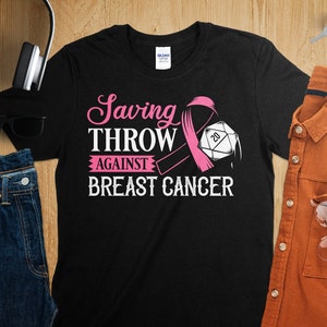 Op de afbeelding: Zwart T-shirt met een roze lint en een dobbelsteen met het nummer 20. De tekst op het shirt luidt "Saving Throw Against Breast Cancer".