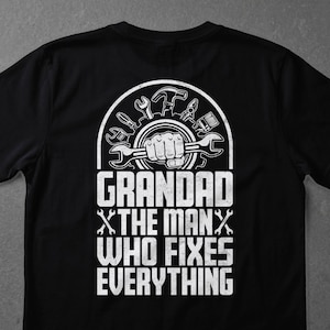 Könnte beinhalten: Schwarzes T-Shirt mit weißer Grafik. Die Grafik zeigt eine Faust, die einen Schraubenschlüssel hält, umgeben von Werkzeugen, und den Text "GRANDAD X THE MAN X WHO FIXES EVERYTHING."