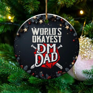 May include: A round Christmas ornament with the text "WORLD'S OKAYEST DM DAD" in white and red. The ornament has a black background with decorative stars, berries, and pine needles around the edge. It is hanging on a Christmas tree.