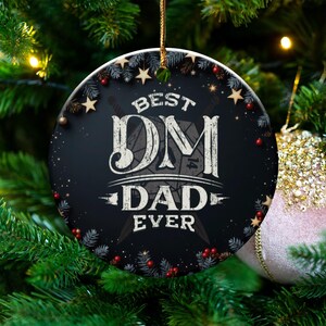 May include: A black and white round Christmas ornament with the text "BEST DM DAD EVER". The ornament is decorated with stars, pine needles, and berries. It hangs on a Christmas tree with a pink ornament.
