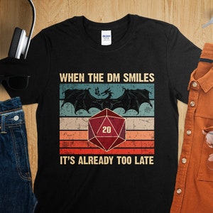 Puede incluir: Camiseta negra con un gráfico de estilo vintage que presenta un dragón, un dado de 20 caras y el texto "When the DM smiles it's already too late".