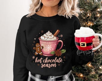 Op de afbeelding: Zwart sweatshirt met een roze afbeelding van warme chocolademelk. De afbeelding bevat een mok met slagroom, een kaneelstokje en een peperkoekmannetje. De tekst "hot chocolate season" is ook aanwezig.
