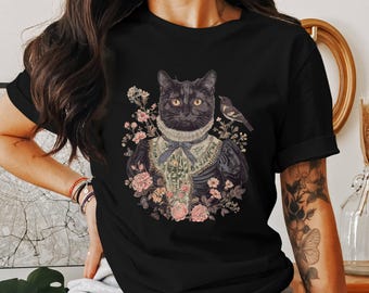 May include: Black t-shirt with a graphic print of a black cat wearing a Victorian-style dress and surrounded by flowers. The cat has green eyes and a white collar. A small bird is perched on a branch above the cat.