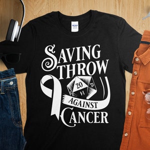 May include: Black t-shirt with a white design of a 20-sided die with a white ribbon and the text "Saving Throw Against Cancer".
