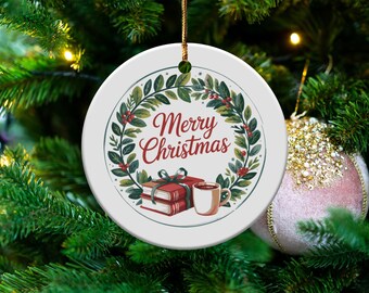 Puede incluir: Adorno navideño de cerámica blanca con un diseño festivo. El adorno presenta una corona con bayas, las palabras "Merry Christmas", una pila de libros y una taza. Cuelga de un cordón dorado.