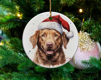 May include: Round Christmas ornament featuring a watercolor painting of a brown dog wearing a Santa hat. The ornament has a white background with red and blue paint splatters. The ornament is hanging on a Christmas tree.