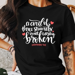 Peut inclure: T-shirt noir avec un texte blanc qui dit "a cord of three strands is not easily broken" et "ecclesiastes 4:12". Le texte est en écriture cursive et il y a trois petits cœurs rouges sur le t-shirt.