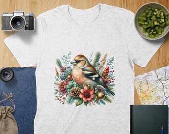 May include: A light gray t-shirt featuring a watercolor illustration of a bird perched amidst flowers, pinecones, and berries. The bird has brown, orange, and black feathers. The design is centered on the shirt, creating a nature-themed aesthetic.