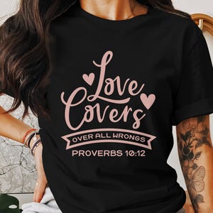 Puede incluir: Camiseta negra con un gráfico rosa que dice "Love Covers Over All Wrongs Proverbs 10:12".