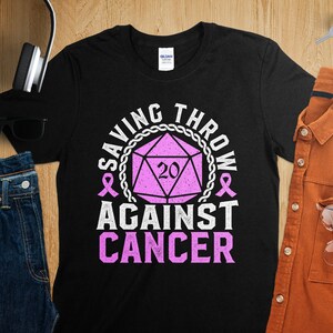May include: Black t-shirt with the text "SAVING THROW AGAINST CANCER" in white and pink. The design includes a pink D20 die and a pink ribbon. The shirt is displayed with other accessories.