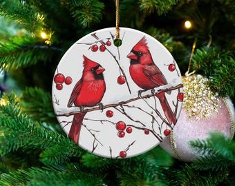 Peut inclure: Ornement de Noël rond représentant deux cardinaux rouges perchés sur une branche enneigée avec des baies rouges. L'ornement a un fond blanc et une ficelle dorée pour l'accrocher. Il est présenté sur un sapin de Noël.