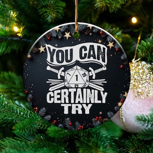Könnte beinhalten: Runder, schwarzer Weihnachtsschmuck mit dem Text "YOU CAN CERTAINLY TRY" in Weiß. Der Schmuck hat einen D20-Würfel, Schwerter und Sterne. Der Schmuck hängt an einem Weihnachtsbaum.