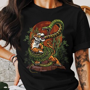 Może przedstawiać: Czarny T-shirt z nadrukiem graficznym przedstawiającym tygrysa atakowanego przez zielonego węża. Wąż jest owinięty wokół ciała tygrysa i ma otwartą paszczę, jakby miał go ugryźć. Tło jest ciemnoczerwone i zielone.