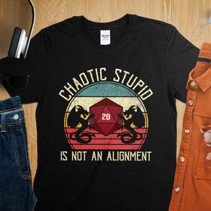 May include: Black t-shirt with a vintage-style graphic of a 20-sided die with a dragon on each side. The text "Chaotic Stupid is not an alignment" is printed below the die.