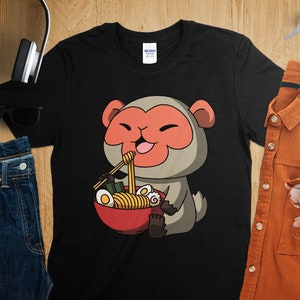 以下が含まれることがあります： ラーメンを食べている漫画の猿が描かれた黒いTシャツ。猿は赤い顔と灰色の体をしており、箸で麺を持っています。卵と海藻が入った赤いラーメンの丼も描かれています。