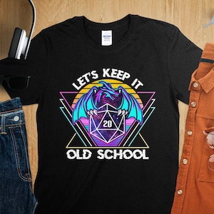 Op de afbeelding: Zwart T-shirt met een retro-grafische afbeelding van een paarse draak die een 20-zijdige dobbelsteen vasthoudt. De tekst "Let's Keep It Old School" staat onder de afbeelding.