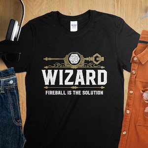 May include: Black t-shirt with a gold design featuring a wizard's staff with a 20-sided die at the top. The text "WIZARD" is in white with the words "FIREBALL IS THE SOLUTION" below.