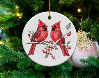 Peut inclure: Ornement de Noël rond avec deux cardinaux rouges perchés sur une branche avec du houx et des baies rouges. L'ornement est blanc avec une ficelle dorée pour l'accrocher. L'arrière-plan est un sapin de Noël.