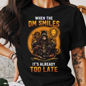 May include: Black t-shirt featuring a graphic of a figure in a hooded robe holding a dice. The text "When the DM Smiles It's Already Too Late" is printed on the shirt.