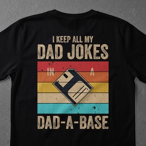 Könnte beinhalten: Schwarzes T-Shirt mit dem Text "I KEEP ALL MY DAD JOKES IN A DAD-A-BASE". Das Design zeigt eine Retro-Grafik einer Diskette auf einem gestreiften Hintergrund in Rot, Orange, Gelb und Blau.