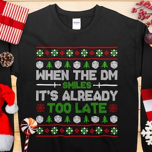 May include: Black sweatshirt with a festive design. The text reads "WHEN THE DM SMILES IT'S ALREADY TOO LATE" in white and green. The design includes snowflakes, trees, and dice, perfect for the holiday season.