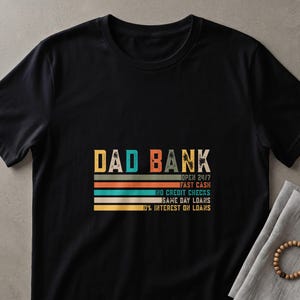 May include: Black t-shirt with the text "DAD BANK" in a retro font. Below the text are horizontal stripes in orange, teal, and yellow. Additional text includes "OPEN 24/7", "FAST CASH", "NO CREDIT CHECKS", "SAME DAY LOANS", and "10% INTEREST ON LOANS".