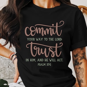 Op de afbeelding: Zwart T-shirt met een roze en witte afbeelding met de tekst "commit your way to the Lord: trust in him, and he will act. Psalm 37:5"
