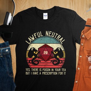 May include: Black t-shirt with a vintage-style graphic of a 20-sided die and a dragon. The text "LAWFUL NEUTRAL" is above the graphic and the text "YES. THERE IS POISON IN YOUR TEA BUT I HAVE A PRESCRIPTION FOR IT" is below the graphic.