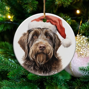 May include: A circular Christmas decoration depicting a brown dog wearing a Santa hat. The ornament has a gold hanging string and is set against a backdrop of a decorated Christmas tree. The dog's portrait is a detailed watercolour.
