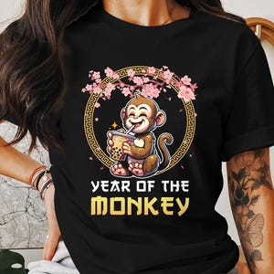 May include: Black t-shirt with a cartoon monkey drinking bubble tea. The monkey is surrounded by pink cherry blossoms and a gold frame with the text "Year of the Monkey".