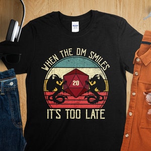 Op de afbeelding: Zwart T-shirt met een vintage-stijl afbeelding van een 20-zijdige dobbelsteen en twee draken. De tekst "When the DM smiles it's too late" is onder de afbeelding gedrukt.