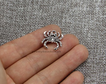 May include: A small, silver spider charm with detailed legs and body segments, and a loop for attaching to jewellery or crafts. The charm is shown against a textured grey background. The text "prettybeadscraft" is visible.