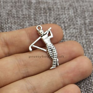 May include: A silver-colored charm depicting a golfer in mid-swing. The charm has a loop at the top for attaching to a bracelet or necklace. The golfer is wearing a checkered skirt. The charm is a miniature golf-themed accessory.