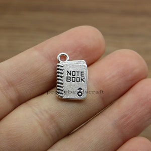 May include: Silver charm in the shape of a notebook with the words "NOTE BOOK" on the front. The charm has a spiral binding detail.